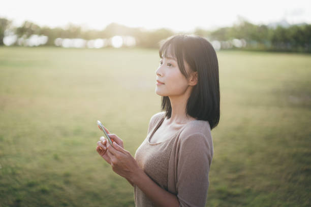 Carefree and serene young Asian woman using smartphone while relaxing in the park and enjoying the sunlight  smart phone green background stock pictures, royalty-free photos & images
