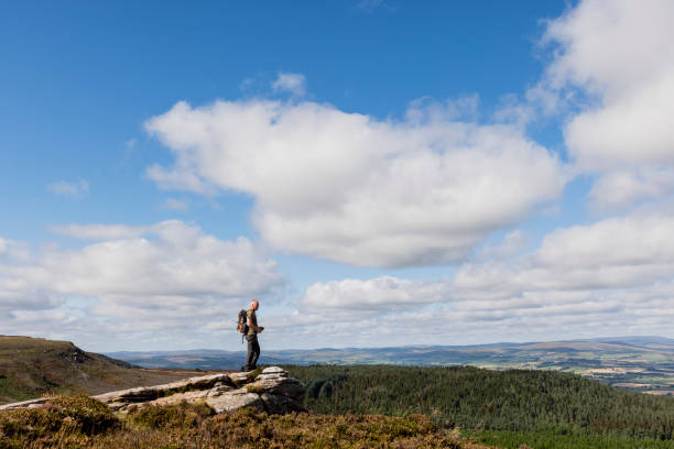 Carefree and Tranquil A senior man wearing a backpack on a hike in the hills in Rothbury, Northumberland. He is standing on a cliff edge and looking at the beautiful green scenery. rothbury northumberland stock pictures, royalty-free photos & images