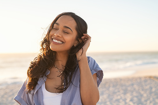 Smiling latin hispanic woman relaxing on beach with closed eyes at sunset. Beautiful mixed race woman enjoying wind fluttering hair. Charming and calm young woman breathing fresh air at summer beach with copy space.