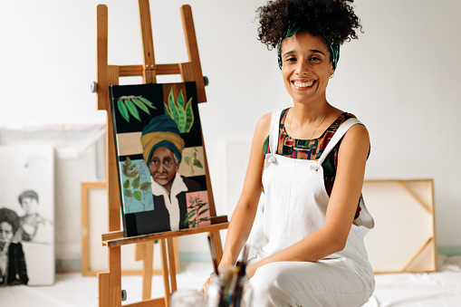 Carefree female painter smiling in her studio. Happy young painter looking at the camera while sitting next to her artwork. Cheerful young woman working on a new painting in her art studio.