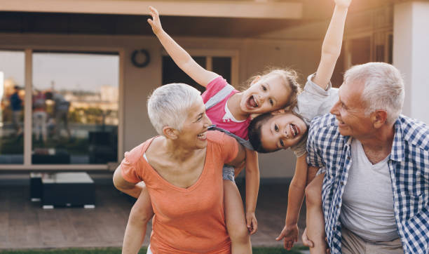Carefree grandparents piggybacking their joyful grandkids in the front yard. Playful grandparents having fun with their grandkids while piggybacking them in the backyard. Children are looking at camera. front yard stock pictures, royalty-free photos & images