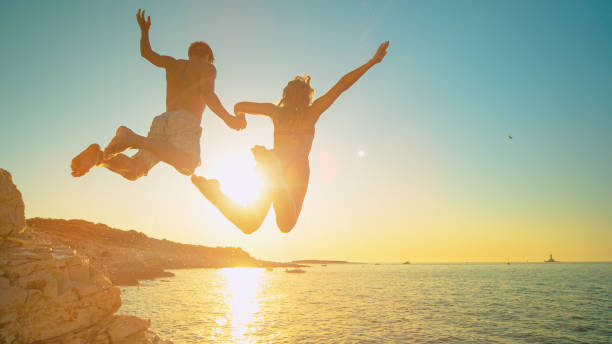 LENS FLARE: Carefree tourists hold hands while jumping into sea at sunset. stock photo