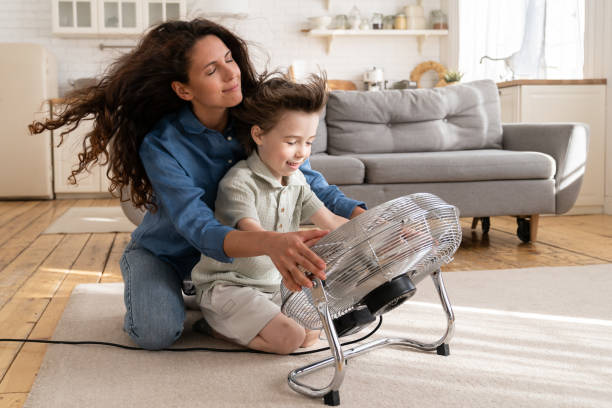 Carefree young mom or nanny have fun with boy son at home sitting together at big fan blowing air stock photo