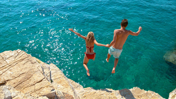 Carefree young tourists holds hands while jumping into the refreshing blue sea. stock photo