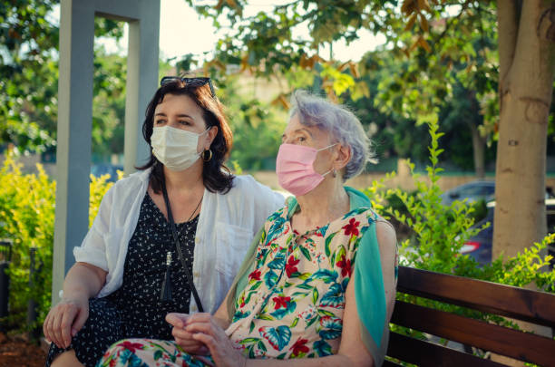 Caregiver and the old woman, wearing masks, sat down to rest on a park bench near the house. stock photo