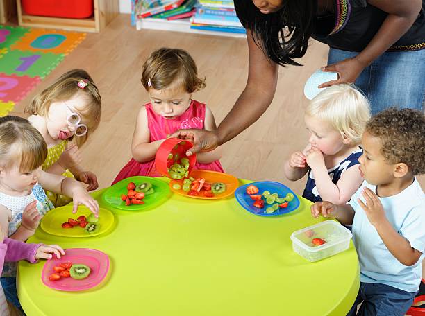 Carer Serving Fruit To Group Of Toddlers/ Children At Nursery A portrait of a carer serving fruit to a group of toddler/ children in a nursery setting. meal of kindergarten stock pictures, royalty-free photos & images