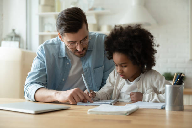 Caring dad help biracial daughter with homework at home Caring Caucasian father help biracial little daughter with homework at home, loving European dad and small African American girl child study together in kitchen on quarantine, homeschooling concept homework stock pictures, royalty-free photos & images