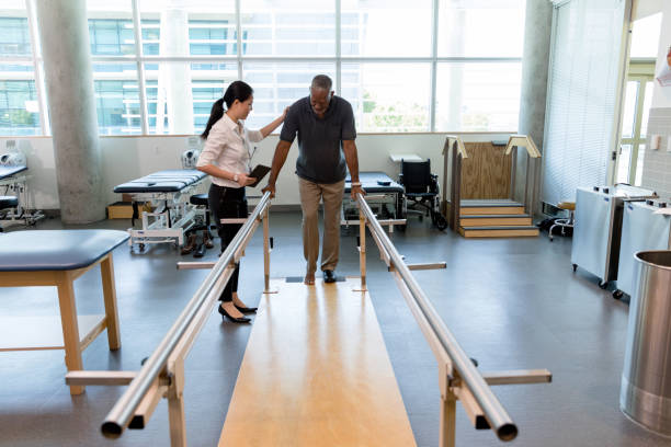 Caring female physical therapist helps stroke victim in rehab center Female physical therapist helps a senior man walk following a stroke. The man is using parallel bars in a rehab center. recovery stock pictures, royalty-free photos & images