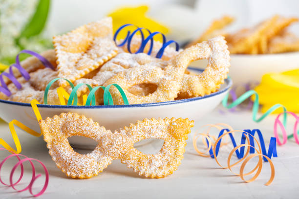 Carnival mask shape Angel wings or sfrappole or chiacchiere. Traditional sweet crisp pastry deep-fried and sprinkled with powdered sugar. Italian carnival food tradition. Decorated with paper serpentine. stock photo