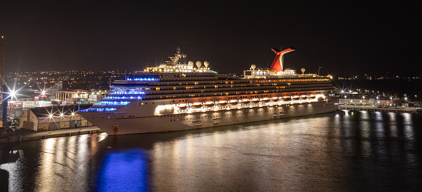 Bridgetown, Barbados - May 1, 2020: Carnival Valor docked in the port of Bridgetown at night. Beautiful reflections on the water in the foreground, port and city lights in the background.