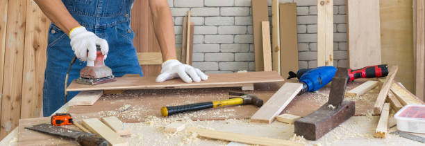 Carpentry use a sander machine to smooth plywood surfaces by abrasion with sandpaper at the woodworking facility. A desk full of hand tools and wood piles. stock photo
