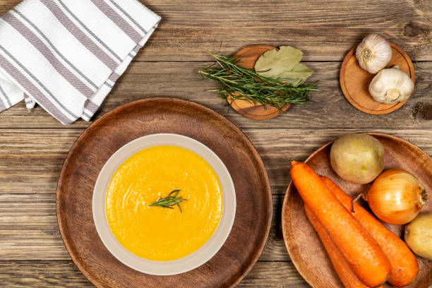 Carrot and potato soup bowl on a drift wood table with vegetables in a wood plate, napkin, garlic, laurel and rosemary. Flat lay. stock photo