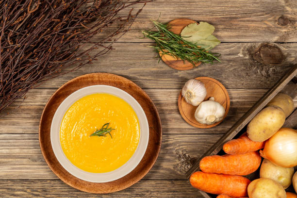 Carrot and potato soup bowl on a drift wood table with vegetables in a crate, dry wood branches, garlic, laurel and rosemary. Flat lay. stock photo