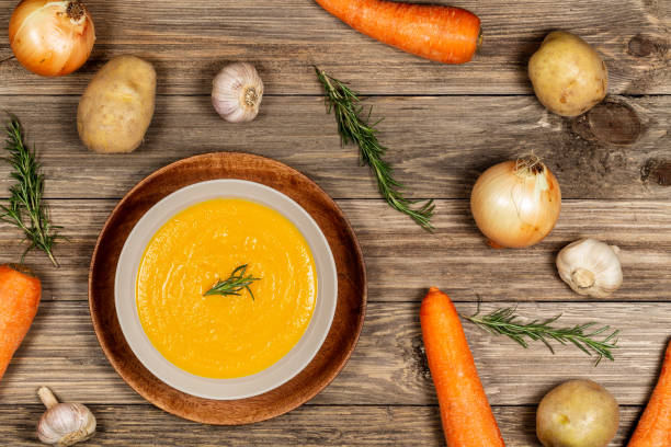 Carrot and potato soup bowl on drift wood table with a spread of vegetables, garlic, and rosemary. Flat lay. stock photo