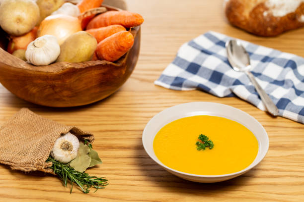 Carrot and potato soup on a wood table with vegetables in a wooden bowl alongside bread, silver spoon, napkin, laurel, garlic and rosemary in a jute bag. stock photo