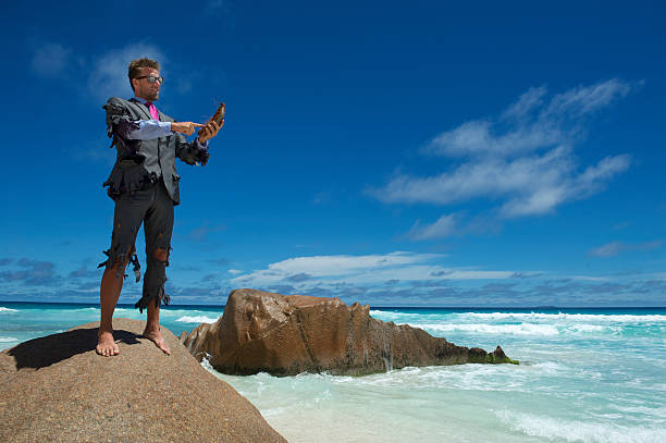 Castaway Businessman Uses Coconut Smartphone on Beach Castaway businessman stands on a rock using his coconut smartphone in a shredded suit desert island stock pictures, royalty-free photos & images