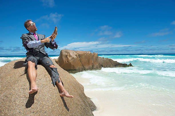 Castaway Businessman Using Coconut Husk Smartphone on Beach Castaway businessman sits perched on a rock using his coconut smartphone in a shredded suit desert island stock pictures, royalty-free photos & images
