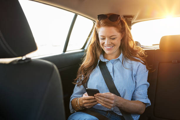 Casual woman using smartphone in car Smiling casual woman sitting in back seat using mobile phone. Cheerful young woman reading messages in smartphone while sitting in a taxi. Attractive girl with red hair wearing blue shirt in car using cellphone. back seat stock pictures, royalty-free photos & images