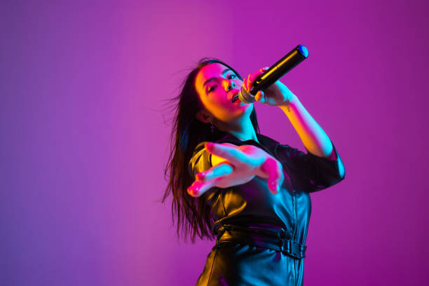 Caucasian female singer portrait isolated on purple studio background in neon light Caucasian female singer portrait isolated on purple studio background in neon light. Beautiful female model in black wear with microphone. Concept of human emotions, facial expression, ad, music, art. singer stock pictures, royalty-free photos & images