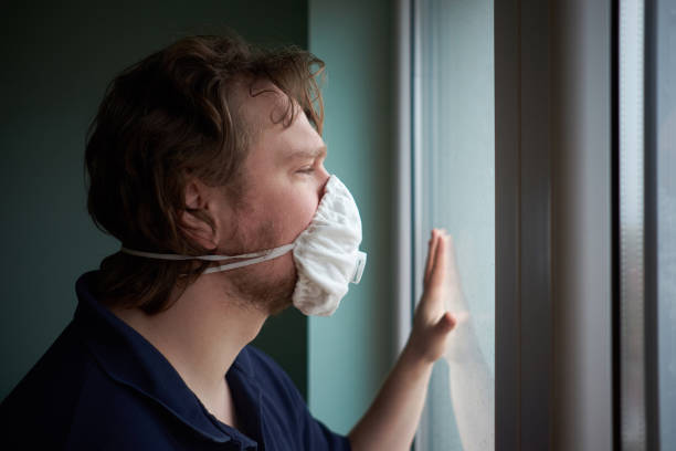 Caucasian man wearing protective medical mask looking out the window during quarantine stock photo