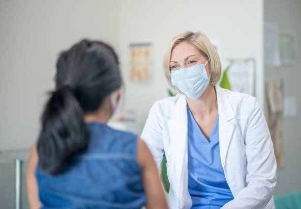 Caucasian medical professional with young female patient 7 year old female patient speaking with her paediatrician in a doctors office, both are wearing masks due to the new COVID-19 regulations and to avoid the transfer of germs. female doctor stock pictures, royalty-free photos & images