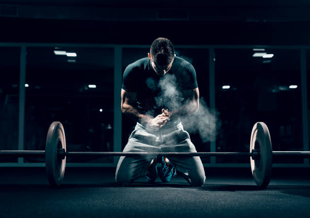 Caucasian muscular man kneeling and clapping hands. In front of him barbell, in background mirror. Gym interior, chalk all around. Caucasian muscular man kneeling and clapping hands. In front of him barbell, in background mirror. Gym interior, chalk all around. face powder photos stock pictures, royalty-free photos & images