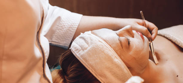 Caucasian spa worker using a brush is applying a skin care cream on the client's face and neck stock photo