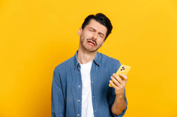 Caucasian stylish guy holding a smartphone in his hand, with a negative unhappy facial expression, received a negative message, or an unwelcome call, stands on an isolated orange background stock photo