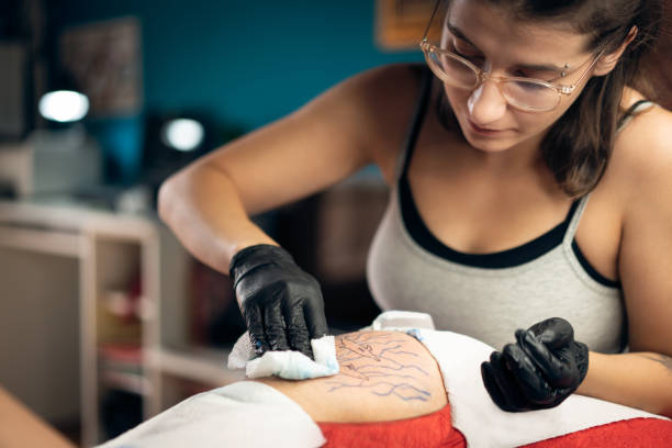caucasian tattoo artist, wiping down the area where she finished with tattooing - tattoo stockfoto's en -beelden