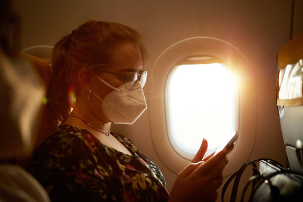 Caucasian Teenage Girl Wearing KN95 Mask on Airplane Side view of 17 year old girl wearing glasses and protective mask checking smart phone while sitting in airplane window seat in time of COVID-19. plane window seat stock pictures, royalty-free photos & images