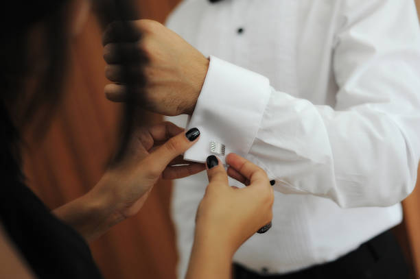 Caucasian woman helping business man or groom wearing vintage silver cuff links on white shirt while getting dressed stock photo