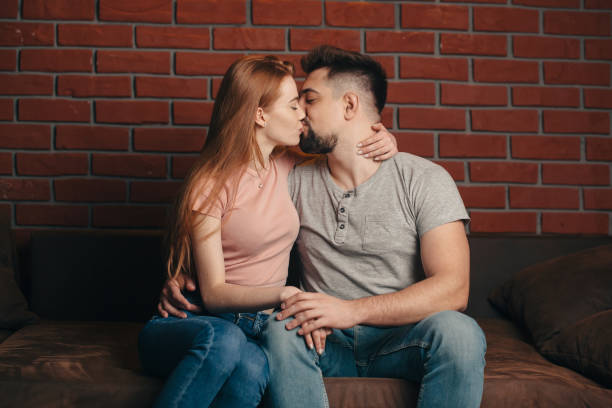 Caucasian woman with red hair and freckles is smiling embracing her lover kissing on a couch at home stock photo