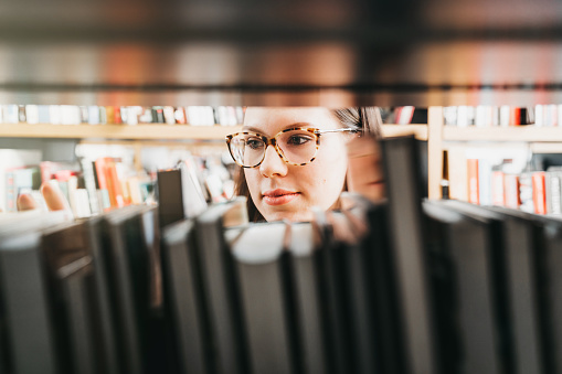 A student girl wearing brown glasses caught in a library, lost between the books on shelves, looking at the books. Picture taken behind the books.