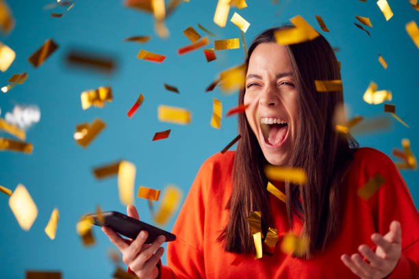 firar ung kvinna med mobiltelefon vinnande pris och duschade med guld konfetti i studio - vinna bildbanksfoton och bilder