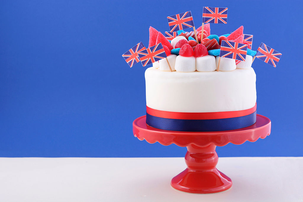UK celebration cake with flags, marshmallow and candy decorations on a red cake stand on a white table against a blue background.