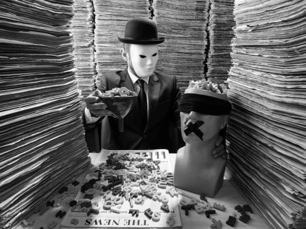 Censor Agent Reading Newspaper In Media Library For Censorship Black and white photo of mid adult man sitting on desk in low key lighting and reading newspaper. Large amount of newspapers are stacked on desk and on the background. He is wearing a black suit, necktie and a bowler hat. His face is obscured with a white mask. A plastic blindfolded mannequin is seen on desk. He is holding a funnel full of letters to fill mannequin brain. The photo was shot with medium format DSLR camera and a wide angle lens in studio lighting. conspiracy stock pictures, royalty-free photos & images