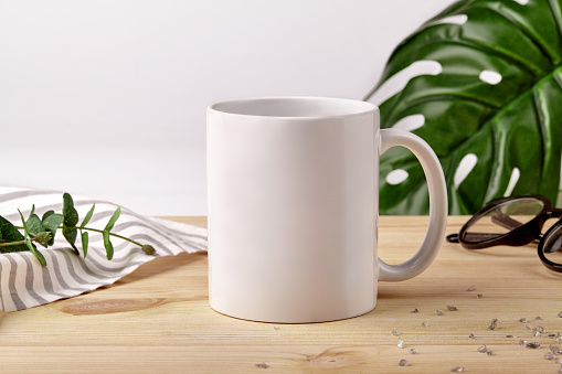 Ceramic mug for coffee or tea on wooden desktop next to striped tablecloth, scattered crystals and green plants against white studio background. Mock up, branding area. Close up, copy space