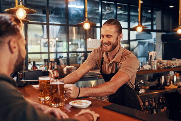 Charming young bartender in apron serving beer and smiling Charming young bartender in apron serving beer and smiling while working in the pub bartender stock pictures, royalty-free photos & images