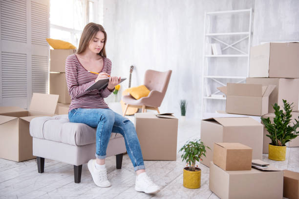 Charming young woman checking list of boxes Everything in order. Beautiful young woman sitting on the pouf in her new flat and checking the list of boxes in the notebook Dorm Essentials stock pictures, royalty-free photos & images