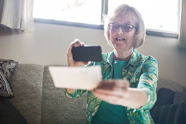 Check Remote Deposit Capture by Senior A smiling senior woman in her 60's takes a picture with her smart phone of a check or paycheck for digital electronic depositing, also known as "Remote Deposit Capture".  She sits in comfort of her home living room.  Horizontal image. bank deposit slip stock pictures, royalty-free photos & images