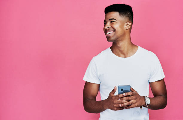 Checking my social media Cropped shot of a handsome young man standing alone and using his cellphone against a pink background in the studio one young man only stock pictures, royalty-free photos & images