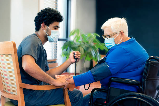 CNA checking patient's blood pressure Portrait of a smling African-American male nursing employee wearing gray scrubs and face mask interacts with a senior aged patient during the Covid-19 pandemic, Midwest, USA elderly care stock pictures, royalty-free photos & images