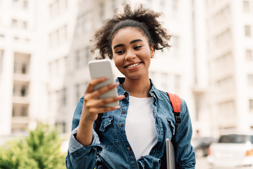 Cheerful African American Student Girl Using Mobile Phone Texting And Learning In Educational Application Standing Outside Near College Building. New App For Your Cellphone Concept