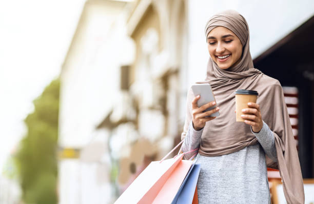 Cheerful arab girl in hijab with shopping bags using phone Cheerful arab girl in hijab with shopping bags and cup of coffee using mobile phone, spending weekend at shopping mall, empty space hot arab woman stock pictures, royalty-free photos & images
