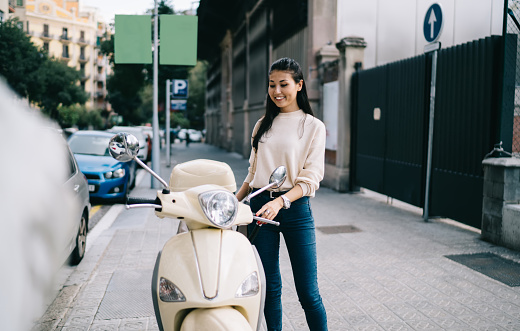 Cheerful Asian female traveller dressed in casual clothing smiling near rented vintage moped enjoying time for getaway driving, happy hipster tourist 20 years old laughing near retro motorcycle