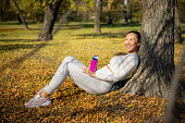 istock Cheerful Asian woman sitting on ground leaning on a tree and facing the sunshine, holding water bottle 1404188789