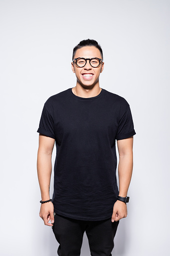 Portrait of handsome asian young man wearing black t-shirt, pants and eyeglasses, laughing at camera. Studio shot, grey background.