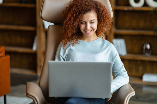 Cheerful attractive businesswoman looks at the laptop screen and smiles Cheerful attractive businesswoman looks at the laptop screen and smiles, happy female freelancer sitting on the armchair and typing, chatting online, enjoying remote work free jpeg images stock pictures, royalty-free photos & images