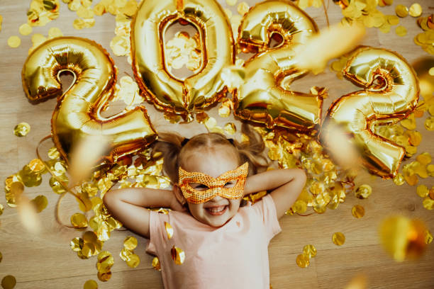 Cheerful Baby girl in mask with numbers 2022 rejoices at the confetti flying from above stock photo
