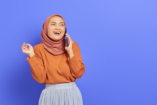 Cheerful beautiful Asian woman in brown sweater and hijab talking on mobile phone while looking aside isolated over purple background. People islam religious concept